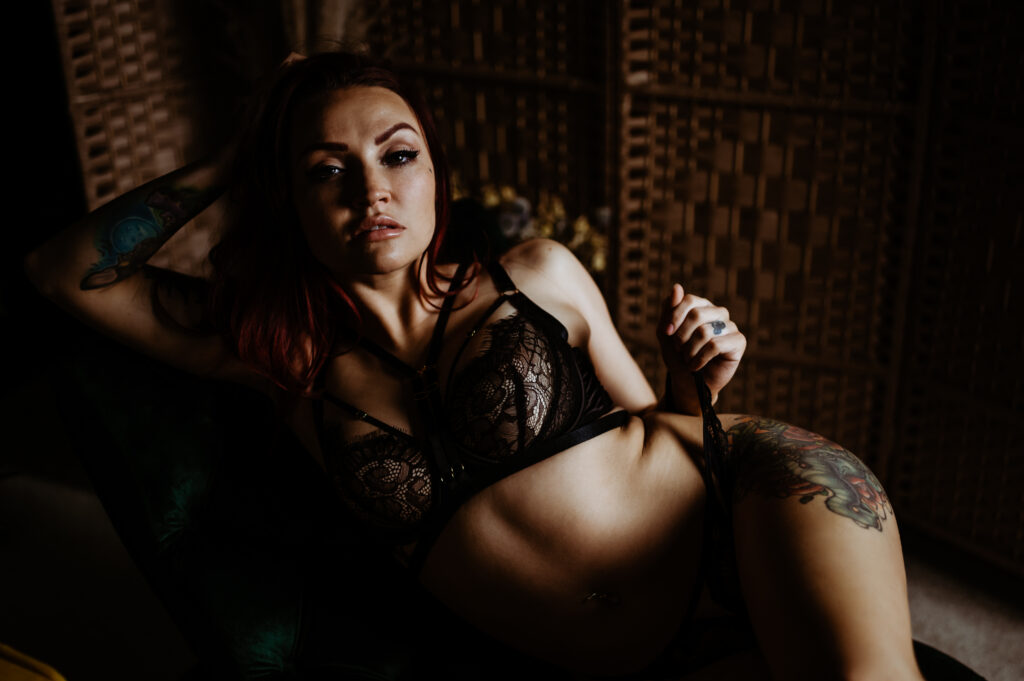 A woman with tattoos poses on a dark green chair, wearing black lace lingerie. She is reclining, looking sexy and confident, with one arm behind her head. The low, dramatic lighting adds a sultry touch to this boudoir photography scene.