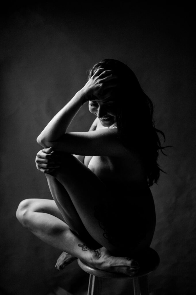 A woman sits nude on a stool in a dark, moody setting, striking a classic boudoir pose—with one arm over her knees and the other hand resting on her head. Soft light beautifully highlights her silhouette, capturing the allure of boudoir photography.
