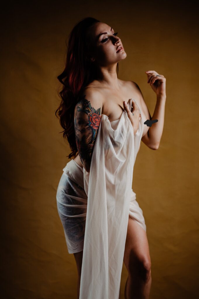 A woman with long dark hair and a colorful arm tattoo poses against a brown background, draped in sheer white fabric. She exudes boudoir photography vibes, gazing upward with a confident expression while looking sexy and elegant.