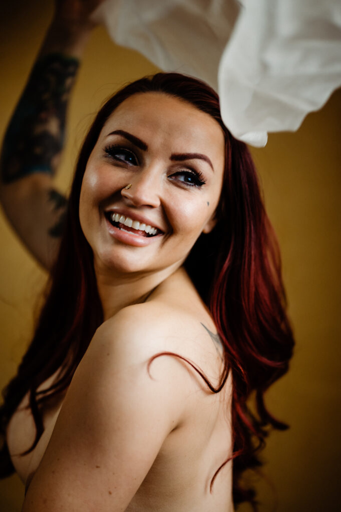 A woman with long red hair and a nose ring smiles brightly while looking to the side, her arm raised. She has a tattoo on her arm and holds up white fabric in a warm yellow setting, capturing the playful elegance of boudoir photography.