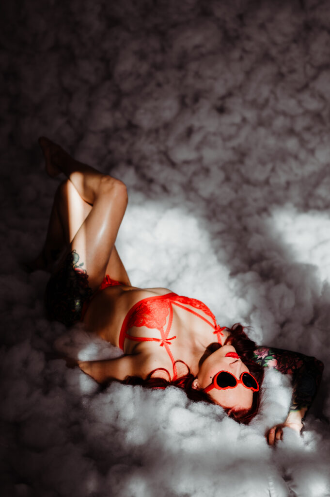 A woman looking sexy in red lingerie and heart-shaped sunglasses lies on a fluffy, cloud-like surface, the sunlight casting soft highlights—a playful boudoir scene.