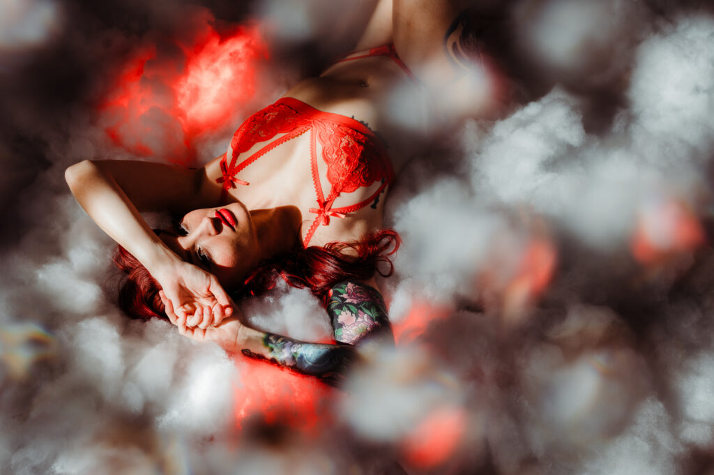A woman with long red hair, wearing a red lace bra and underwear, lies on her back surrounded by soft clouds or smoke in a boudoir photography setting, looking sexy with one arm resting on her forehead and the other beside her head.