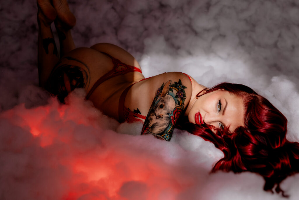 A woman with red hair, red lipstick, and colorful tattoos poses looking sexy in red lingerie, lying on her stomach amidst fluffy white clouds with a glow of red light beneath her—a stunning moment of boudoir photography.