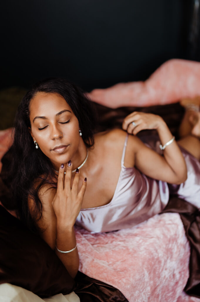 A woman in a silky lavender dress reclines on a bed with pink and brown bedding, eyes closed and hand gently touching her neck, exuding a serene and relaxed mood—a beautiful moment of boudoir photography, effortlessly looking sexy.