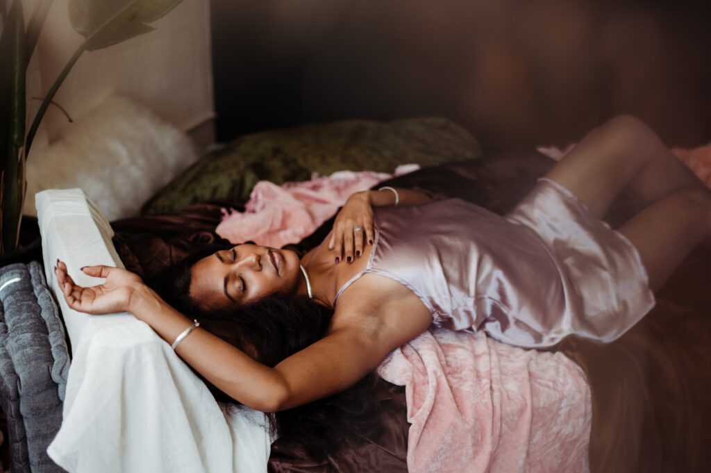 A woman in a silky lavender nightgown lies on a bed covered with pink and dark blankets, exuding boudoir elegance as she rests with her eyes closed and one arm above her head, appearing peaceful and relaxed.