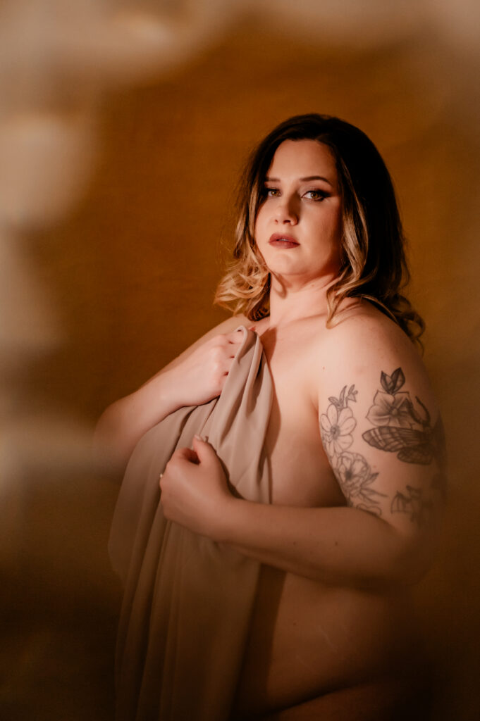 A woman with wavy hair poses nude, partially covered by a beige cloth. In this boudoir photography scene, she has a floral tattoo on her upper arm and gazes confidently at the camera against a warm, softly blurred background.