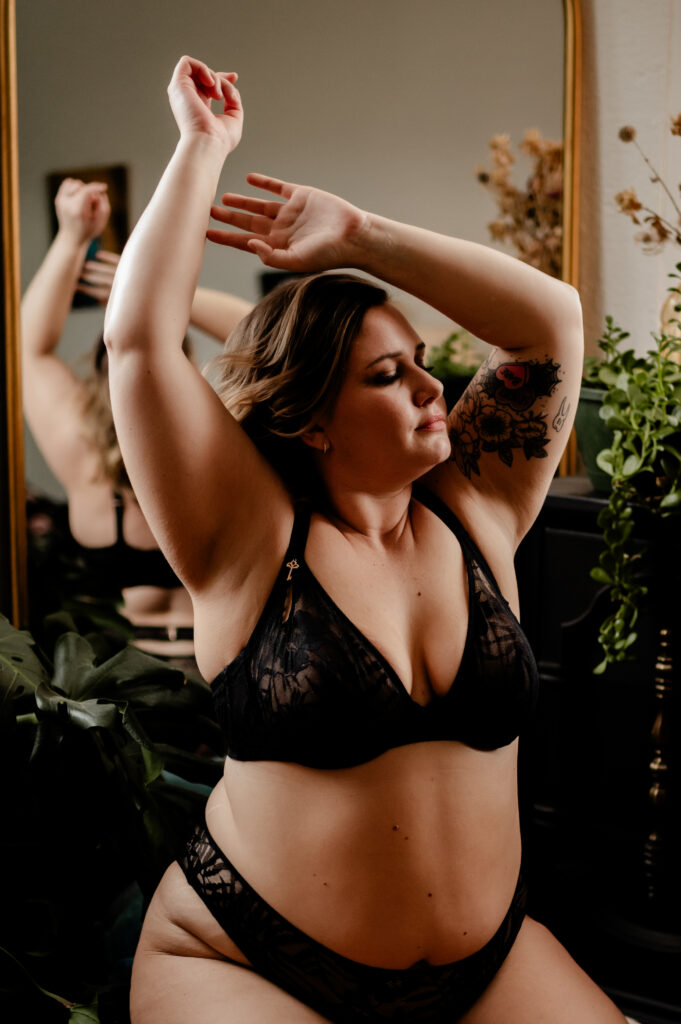 A woman looking sexy in black lace lingerie poses with her arms raised in front of a mirror. She has a floral tattoo on her arm and is surrounded by indoor plants, with soft, natural boudoir lighting highlighting her features.
