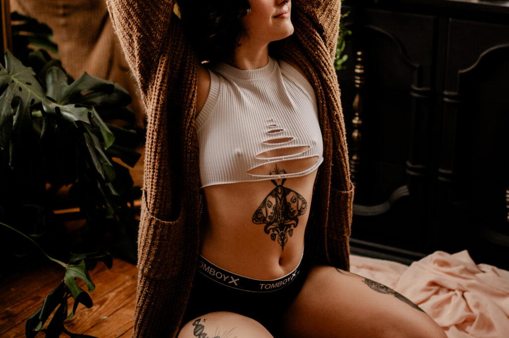 A person looking sexy in a white ripped crop top, black underwear, and a brown cardigan sits with arms raised. Visible tattoos adorn their abdomen and thighs, creating a chic boudoir vibe amid indoor plants and warm-toned decor.