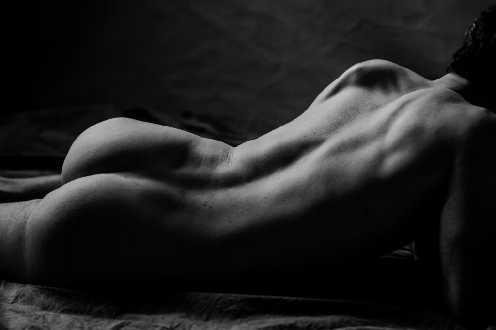 Black and white boudoir photo of a nude person lying face down, showing their bare back, shoulders, and legs, with shadows highlighting muscle definition and contours, capturing the essence of looking sexy.