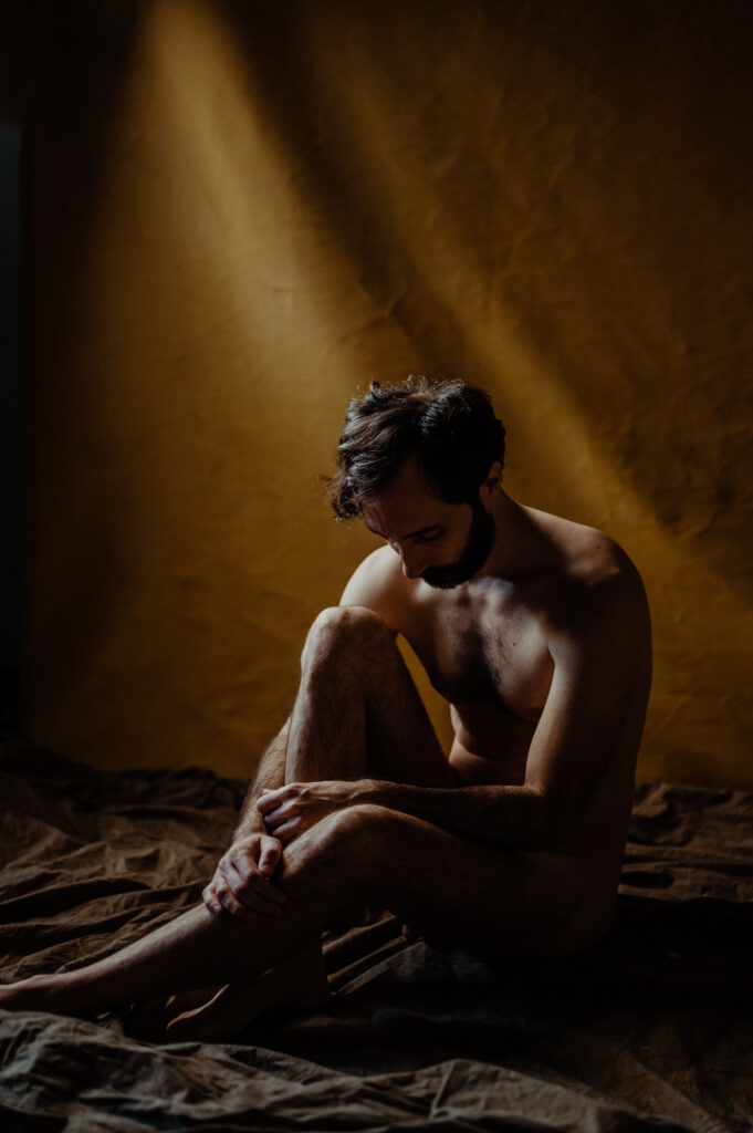 A nude man with dark hair and a beard sits on the floor with knees drawn to his chest, head bowed, and one arm resting on his legs. Warm light highlights him against a brown fabric backdrop in a boudoir photography style.