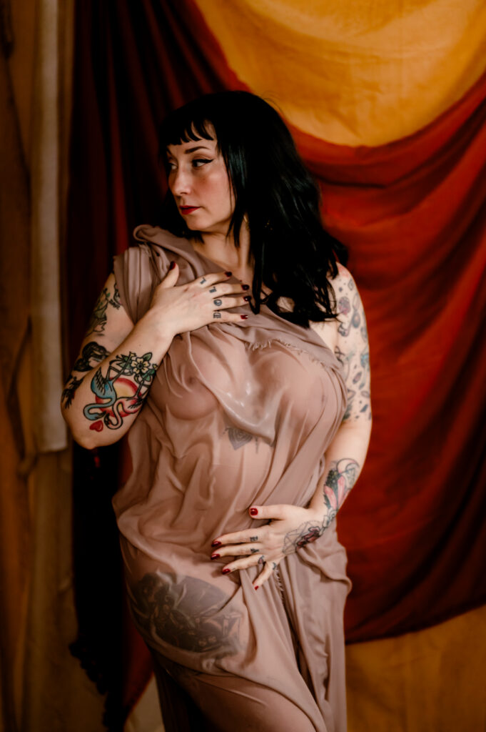A woman with dark hair and tattoos poses in front of draped fabric, wearing a sheer, brownish dress. In this boudoir photography shot, she holds the fabric to her chest and abdomen, looking to the side and exuding a sexy confidence.
