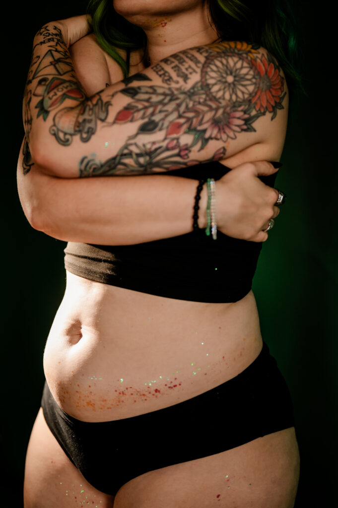A person with colorful tattoos on their arms poses boudoir-style in black underwear and a black top, arms crossed over their chest. Their skin is decorated with scattered glitter, and they wear bracelets on one wrist, confidently looking sexy.