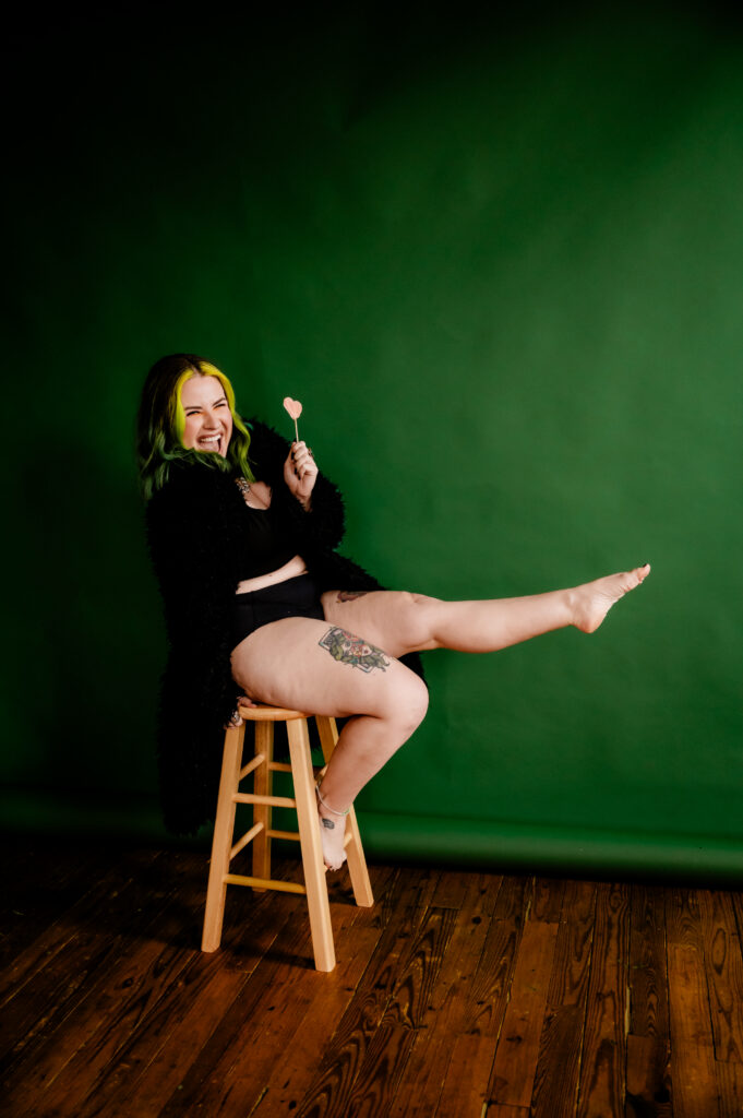 A person with green and yellow hair sits on a wooden stool against a green background, wearing a black outfit and black fur coat, smiling, holding a pink heart lollipop, and playfully kicking one leg up in a fun boudoir-inspired pose.
