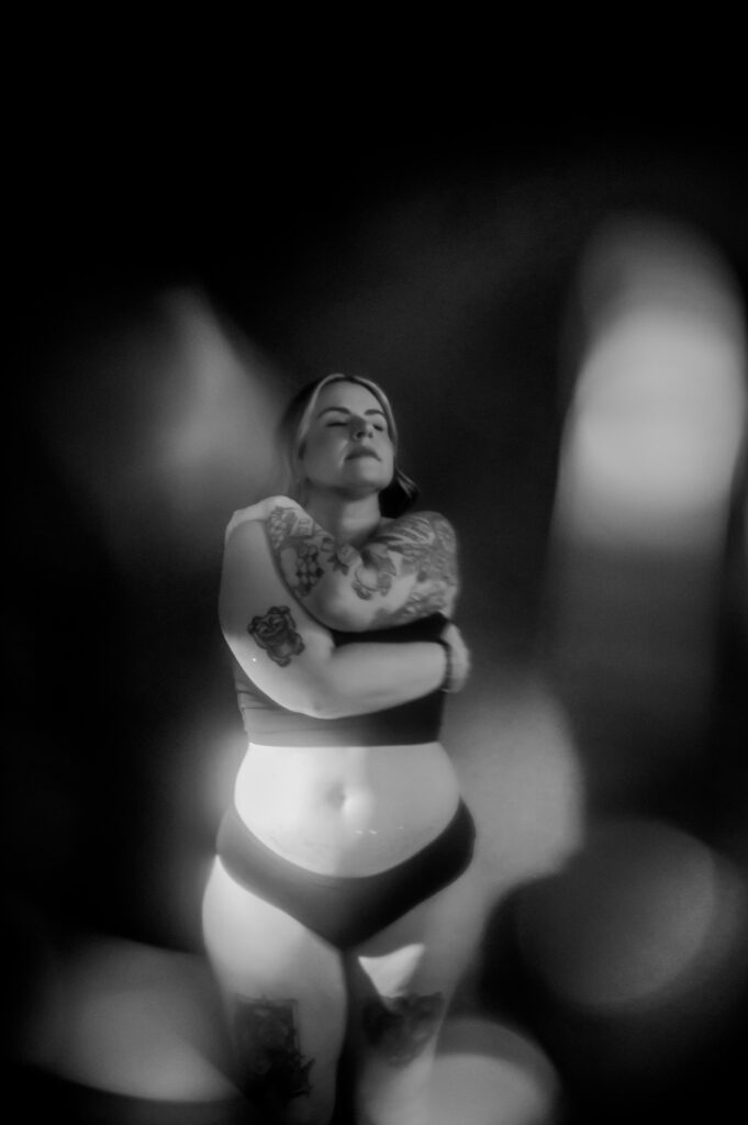 A woman in underwear with visible tattoos stands with eyes closed, arms wrapped around herself in a self-hug. The black and white Boudoir photo features dramatic lighting, soft blur, and shadows, capturing a raw, looking sexy confidence.