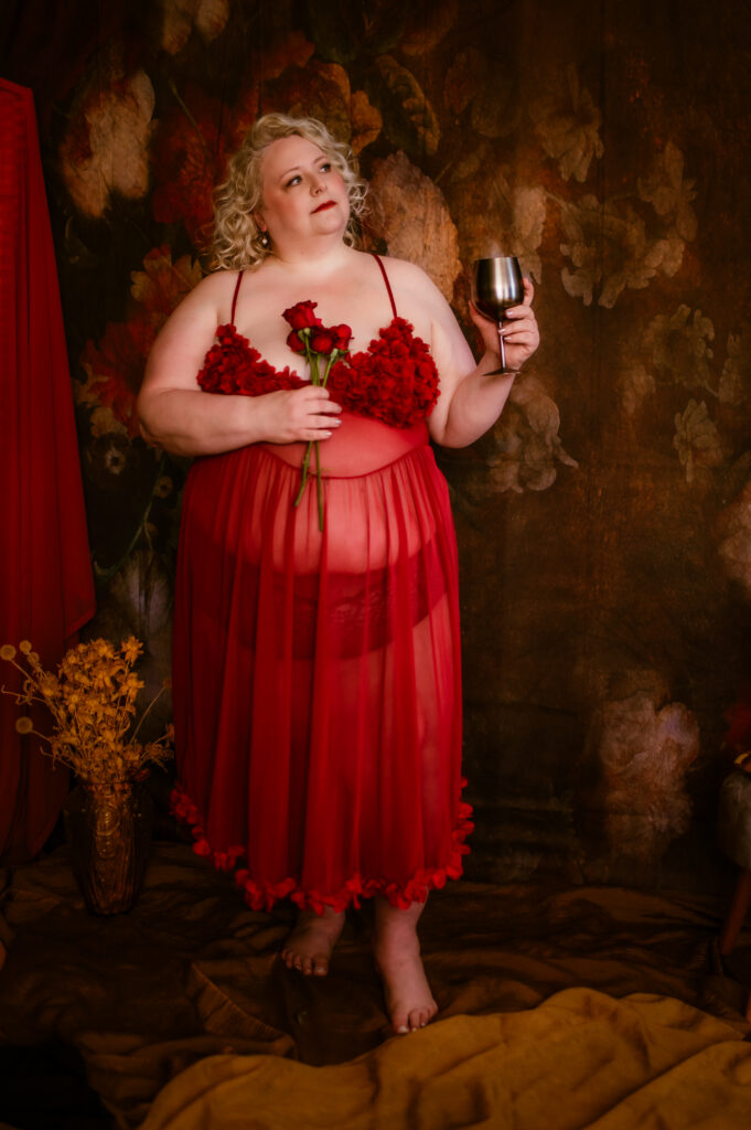 A person with curly blonde hair wears a sheer red dress, looking sexy and confident, holding red roses in one hand and a silver goblet in the other, set against a dark, floral-patterned background—a striking boudoir photography moment.