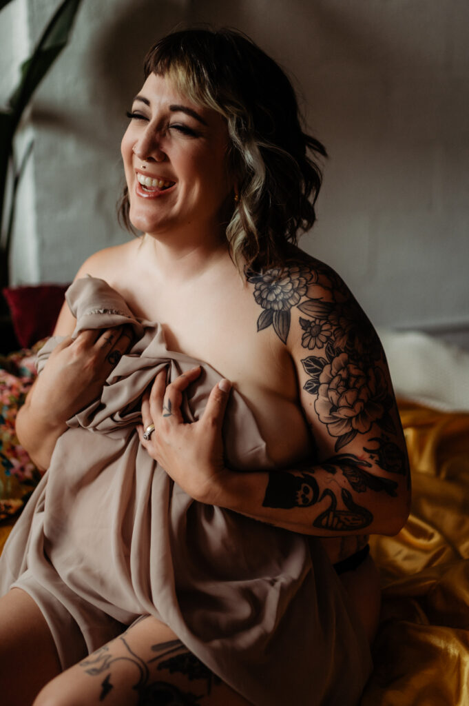 A smiling person with shoulder-length hair and visible tattoos on their arm and leg sits on a bed, holding a beige sheet against their chest. Warm, natural light enhances their joyful expression in this boudoir photography moment.