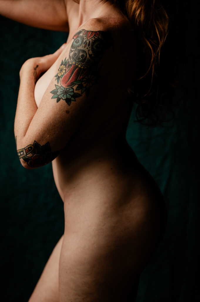 A nude person is shown from the side with one arm crossed over their chest, their upper arm and shoulder adorned with colorful tattoos. The dark background enhances the intimate, boudoir photography style, while their face remains hidden.