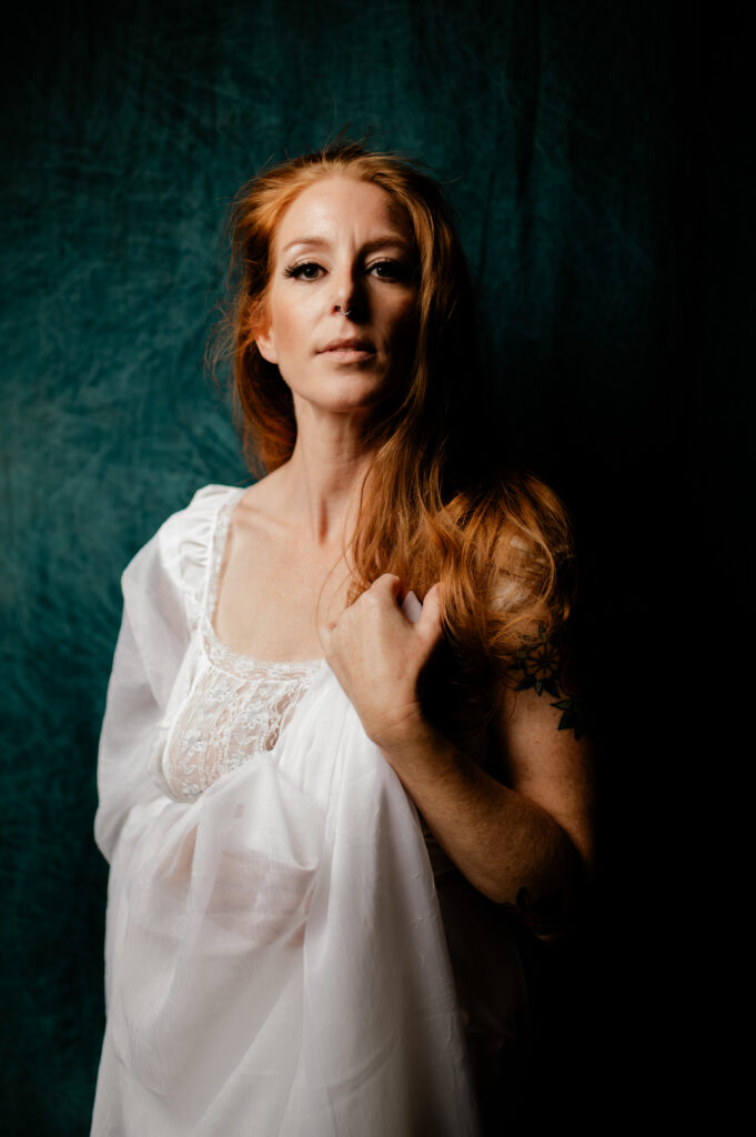 A woman with long red hair stands against a dark green backdrop, wearing a white lace boudoir dress and sheer fabric. She looks confidently at the camera, holding part of the fabric over her shoulder, effortlessly looking sexy.