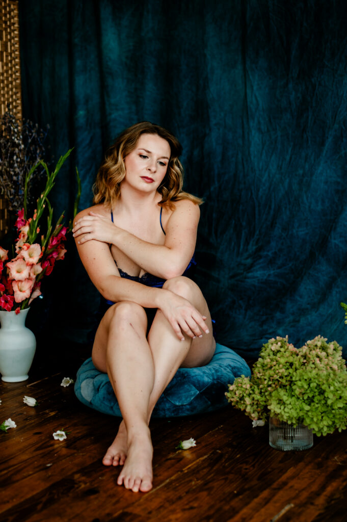 A woman in blue lingerie sits on a cushion with one arm resting on her shoulder, looking sexy amid elegant flower arrangements on a wooden floor, with a blue fabric backdrop enhancing the boudoir photography setting.