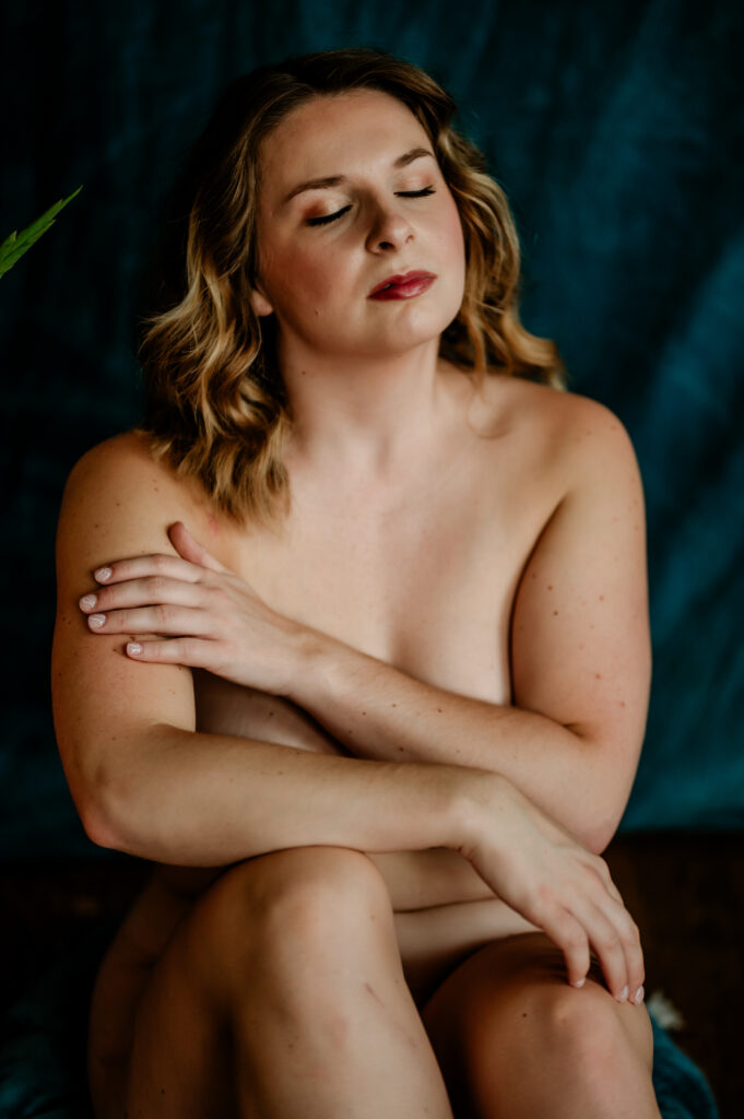 A nude woman with shoulder-length wavy hair sits with her eyes closed, gently resting one hand on her upper arm, capturing the intimate allure of boudoir photography against a dark blue background.