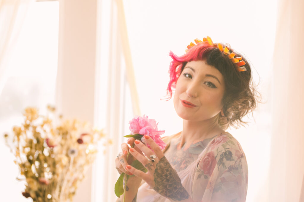A woman with curly hair dyed pink at the front, wearing a floral robe and flower crown, smiles softly while holding a pink flower in a sunlit boudoir filled with dried flowers in the background.
