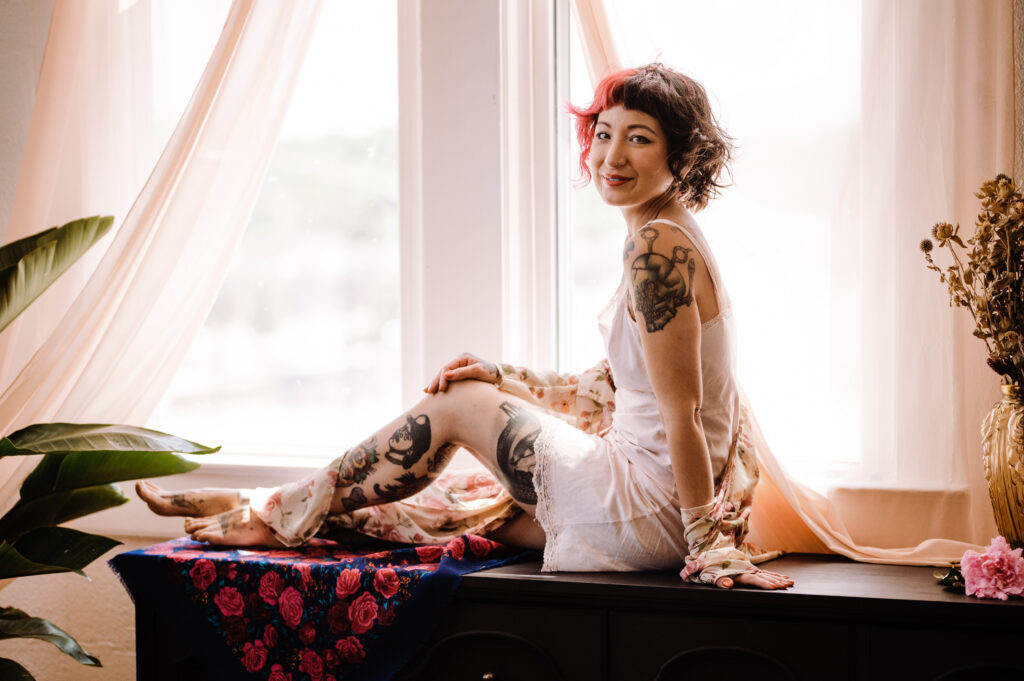 A woman with short brown hair and tattoos sits on a black surface by a large window with sheer curtains. She wears a white dress, smiling at the camera in this intimate boudoir photography scene, surrounded by a floral cloth and dried flowers.