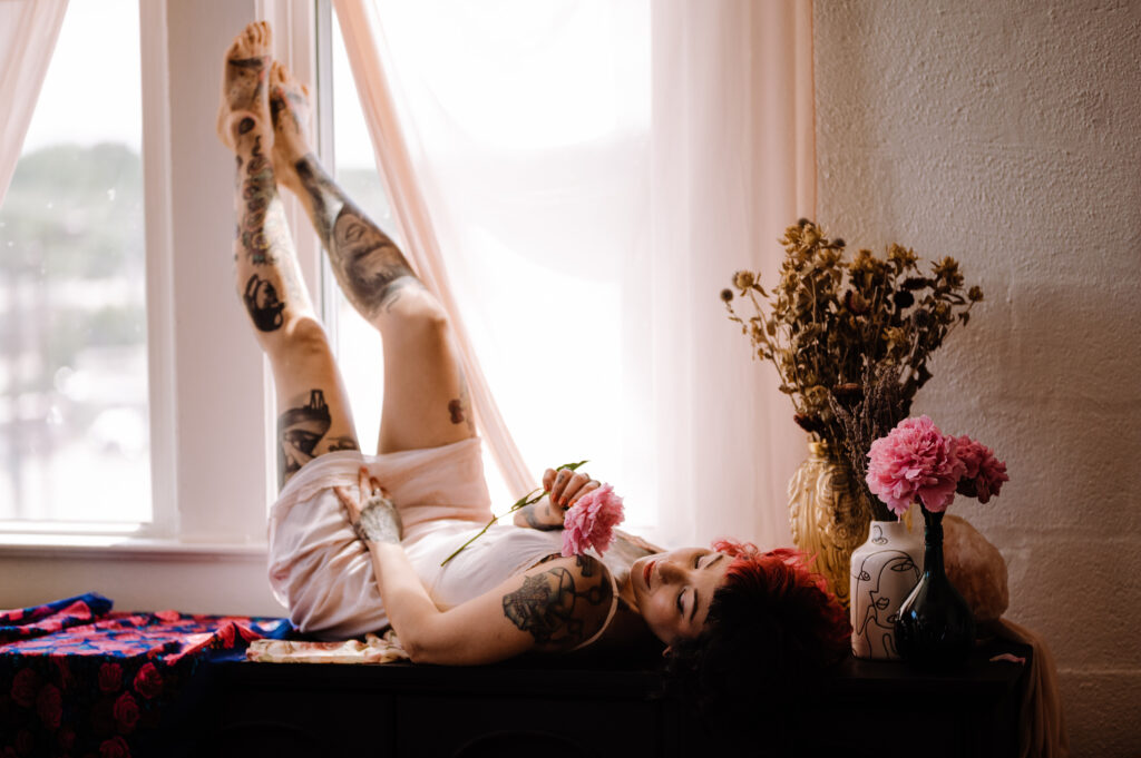 A tattooed person in a white outfit lies on their back on a black cabinet by a window, holding pink flowers. Their legs are raised as soft light filters through sheer curtains—a dreamy boudoir photography scene with dried flowers and a vase nearby.