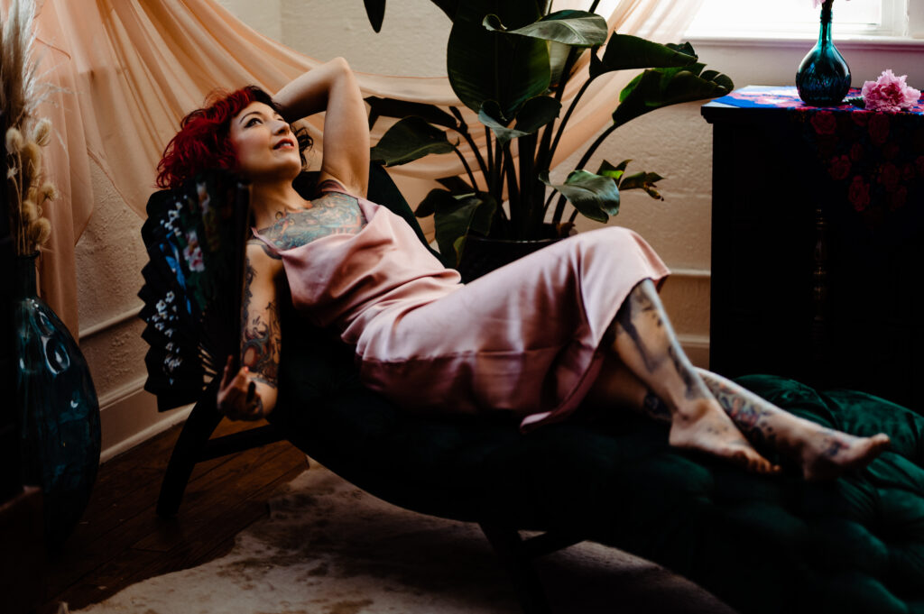A woman with red hair and tattoos lounges on a dark green chaise, wearing a pink satin dress. Striking a boudoir pose, she holds a fan and looks up, surrounded by plants and soft peach drapes in a cozy, softly lit room.