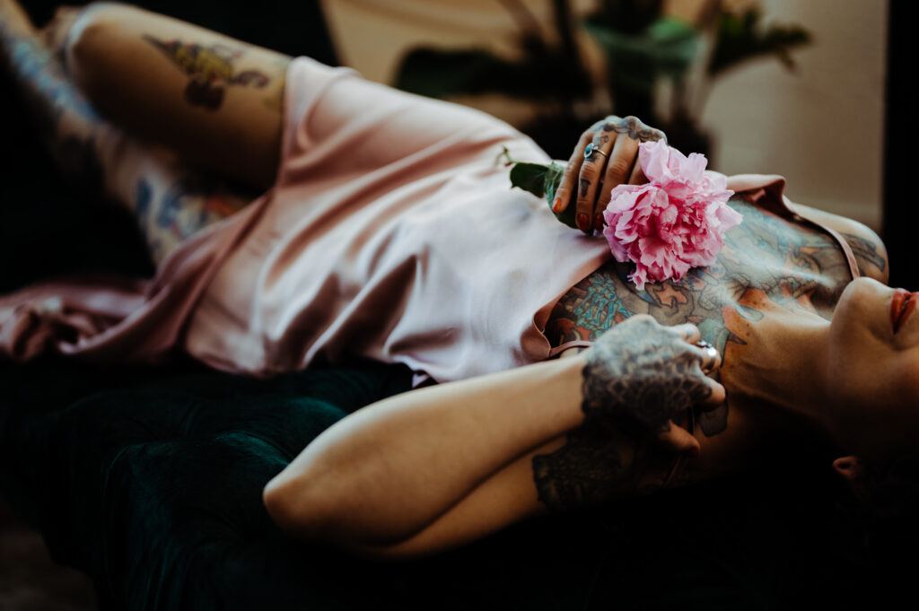 A tattooed person in a silky pink dress lies on a dark green surface, holding a pink peony on their chest. Their head is tilted back in a relaxed pose, capturing the essence of boudoir photography and looking sexy.
