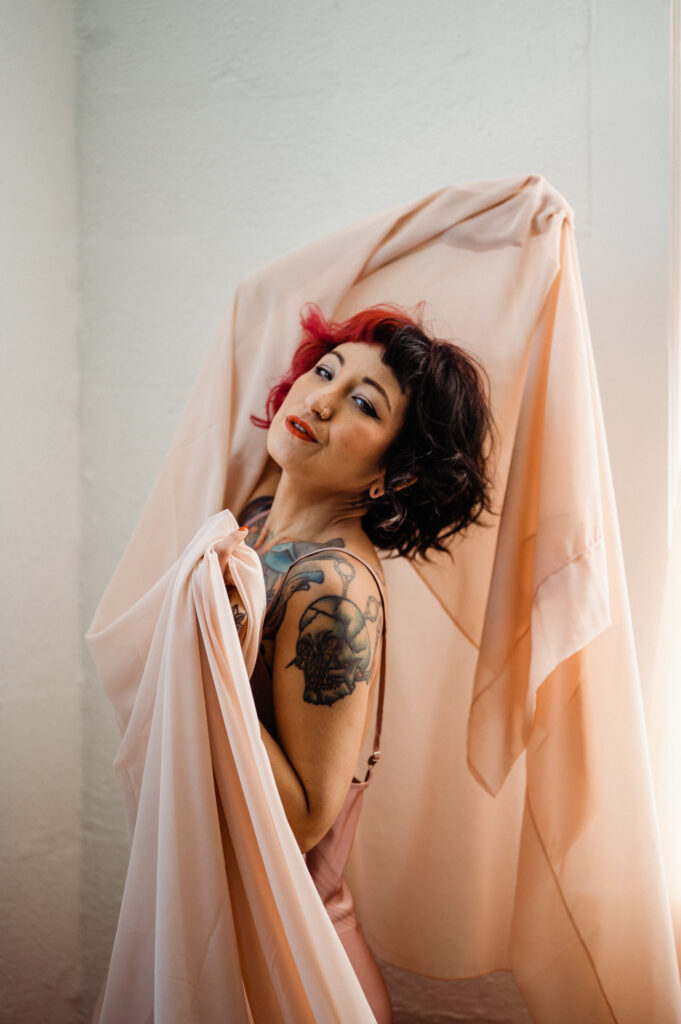 A person with short, dark hair dyed red at the roots poses indoors for boudoir photography, draping a peach-colored fabric. They have visible tattoos on their upper arm and wear bold makeup with red lipstick. Light shines from the right side.