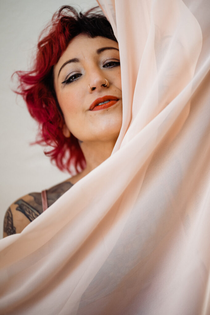 A person with short, curly, red hair and facial piercings smiles softly behind sheer, pale pink fabric, their tattoos visible on their shoulder—capturing a subtle looking sexy vibe reminiscent of boudoir photography.