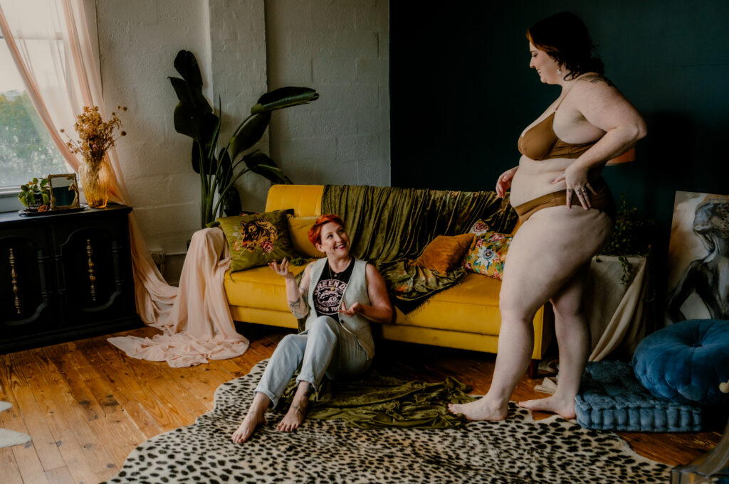 Two women in a cozy, colorful boudoir studio in Nashville, TN; one stands in beige underwear, looking sexy, while the other sits on the floor smiling up at her. The yellow couch, plants, draped fabrics, and natural light create a boudoir-inspired scene.