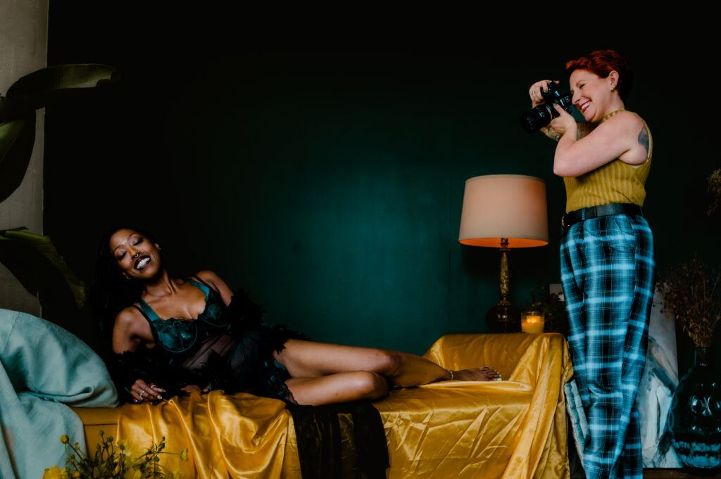 A woman in lingerie reclines on a bed covered with yellow fabric, looking sexy and smiling, while another woman with short hair and plaid pants stands nearby, taking her photo for a nashville boudoir photography session. A lamp and plants are in the background.